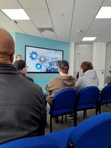 Some engineering students sit facing away from us, towards a screen on a teal-coloured wall, to take in a presentation about Engineering. Just to the side of the screen a presenter can be seen delivering a talk,