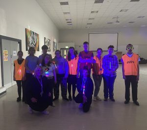 In a large dark room, a group of students pose wearing clothing that is glowing neon colours by ultraviolet light cast at them. Some of the students have long, thin rainbow coloured light pendants hung from their necks. A student on the right has a pair of novelty neon glasses on his face that glow brightly. Most of the group have decorative neon dots painted onto their faces in patterns, glowing in the UV light brightly