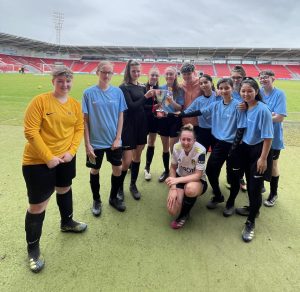 Standing in a close huddle, a group of female football players smile and pose inside Doncaster Rovers stadium, with large banks of red seating in the stands in the distance behind them