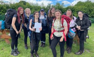 A group of 8 students wearing hiking clothing and large, loaded rucksacks on each of their backs stand smiling toward us. Just behind the group are two picnic tables and further behind is a long green hedgerow. One of the students in the centre holds a piece of paper or map in both hands