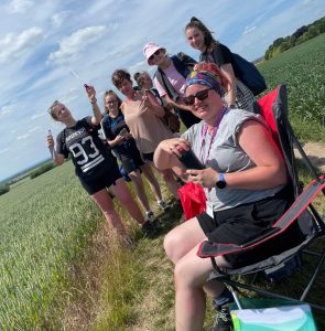 On a bright sunny day on a path surrounded by green fields, a member of staff sits in a camping chair in front of a group of students all dressed for the sunny weather, all of them smiling towards us. The student on the left of the rear group holds a bubble wand in the air creating bubbles around the area