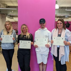 Four Hair and Beauty students stand in our salon side by side, holding certificates and smiling. A bright pink wall is behind them