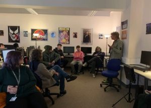 In a classroom with multiple pieces of artwork on the walls, creative media students sit in office chairs around John Parr, musician, who stands and is deep in conversation delivering a talk to the students