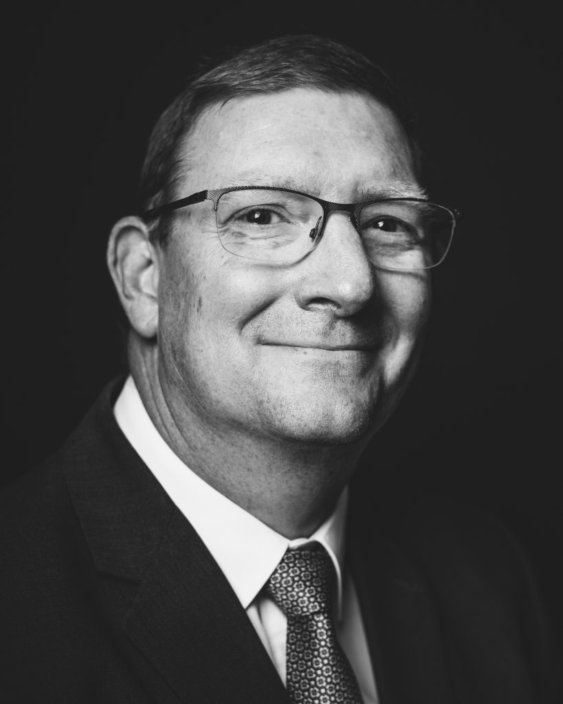 Black and white photograph of John Rees, CEO of DN Colleges Group, smiling and posing in a suit and dark tie