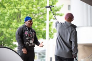 Against a background of a bright green tree, a student is seen talking while being recorded for a video project. Another student closer to us holds a large microphone on a pole