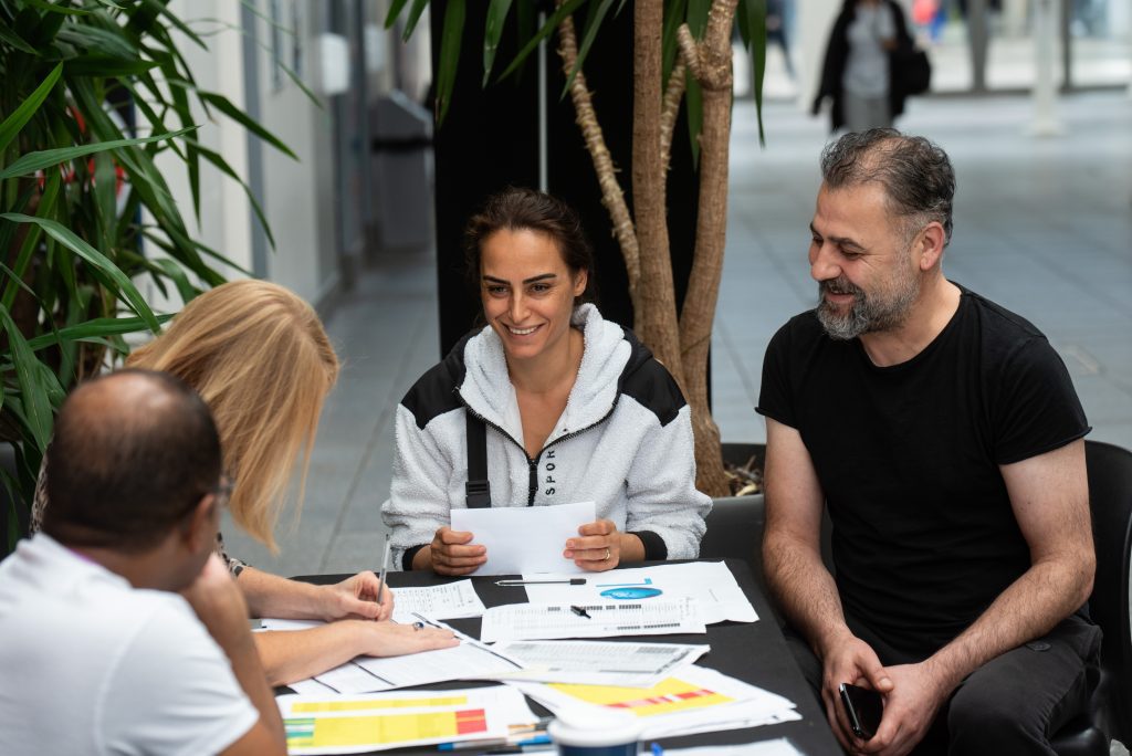 Sat around a table containing flyers and booklets, two people smile while chatting, focussed on two members of staff sat opposite them. In the background, various plants break up the large open space where a few people can be seen walking by.