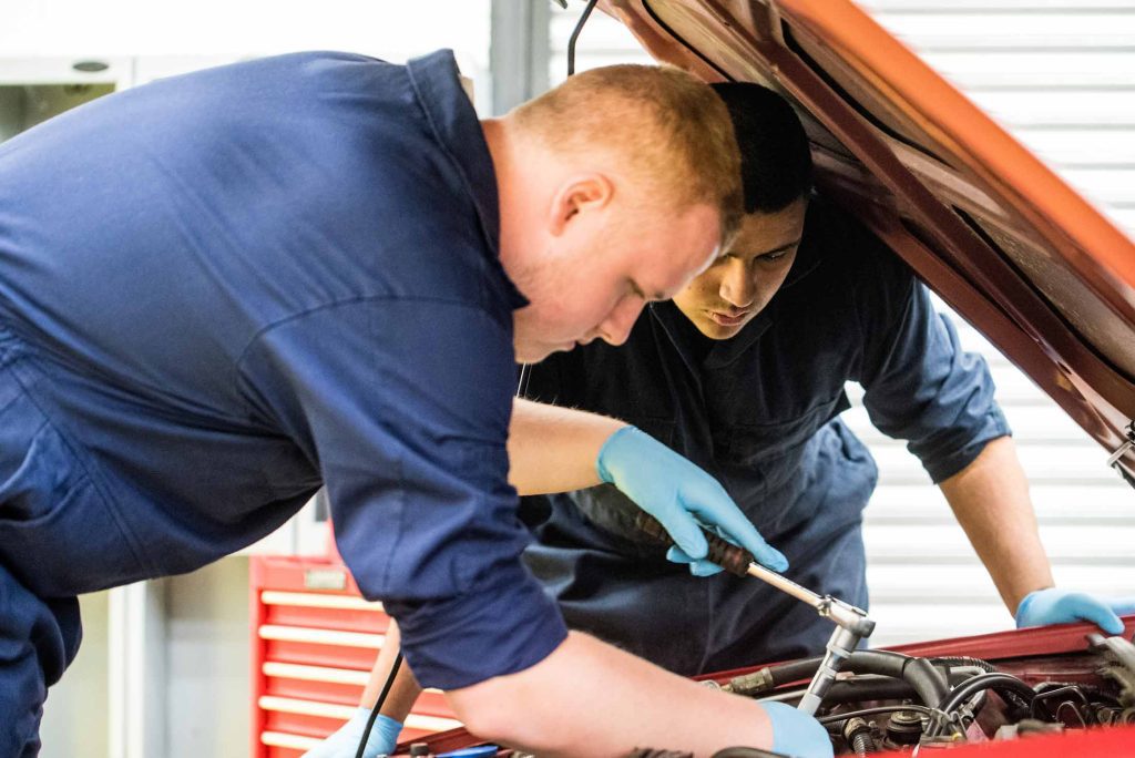 Leaning over into the engine bay of a car, two students look intently at the work they are undertaking. The student nearest to us has a socket spanner placed over a bolt within the engine bay.