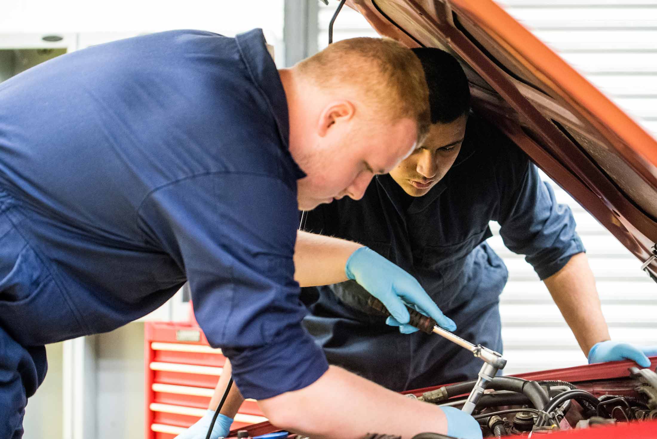 Leaning over into the engine bay of a car, two students look intently at the work they are undertaking. The student nearest to us has a socket spanner placed over a bolt within the engine bay.