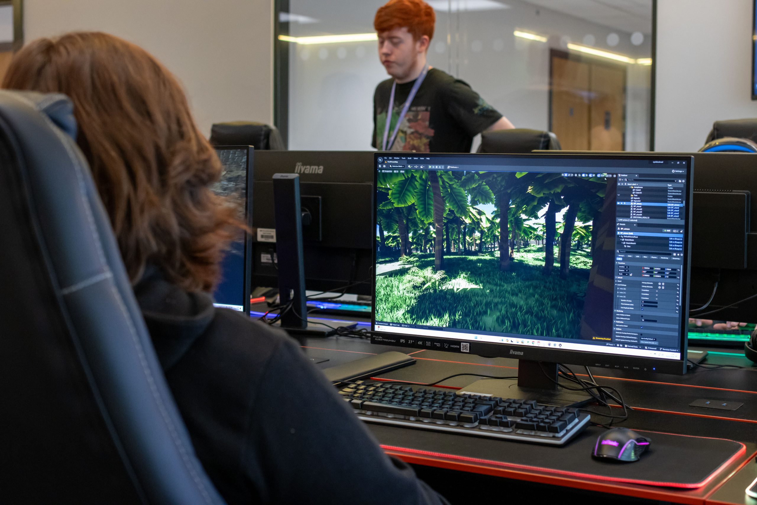 A student sits in a tall gaming chair facing away from us. In front of the student is a computer screen on a bright red trimmed dark desk. On the screen is a program featuring lots of settings and tools, with a big scene showing a view of a 3D rendered tree canopy over a lush green floor.