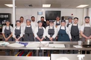 Photograph with 10 students posing in their chef uniforms along side members of staff in a kitchen setting