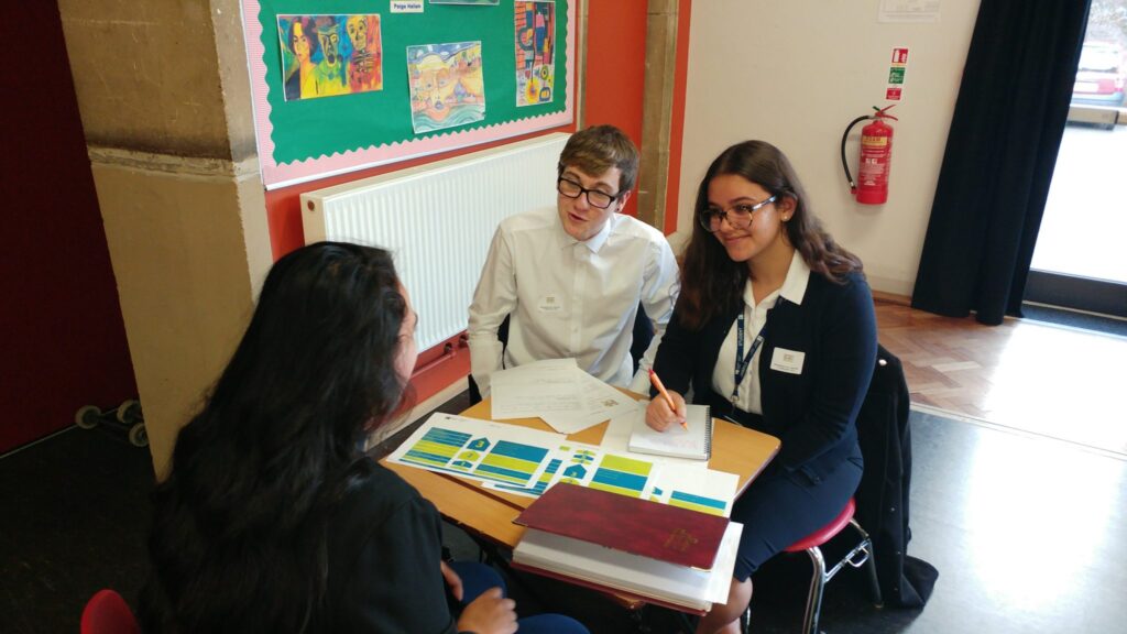 In a classroom setting, 3 students sit around a table, with a few documents on the table between them as they smile and chat to each other.