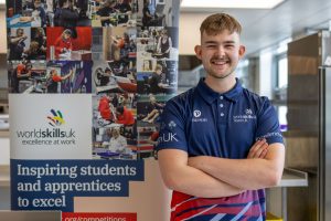 A photo of Oska stood smiling with his arms crossed. He is stood next to a pop-up banner with lots of photos on it and the WorldSkillsUK logo.