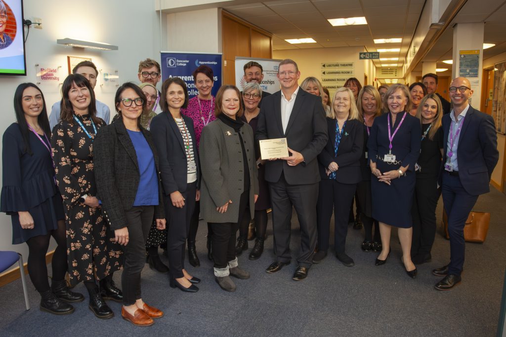 Standing in a large group together, over 20 people pose with happy expressions looking towards the camera. In the centre, DN Colleges Group's John Rees holds a golden plaque presenting it to the group.