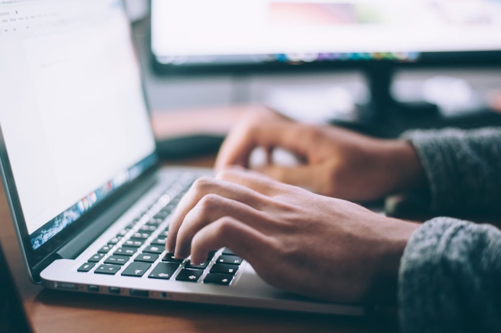 A close up photograph of a person's hands typing at a laptop computer, with the bright screen casting light onto their fingers.