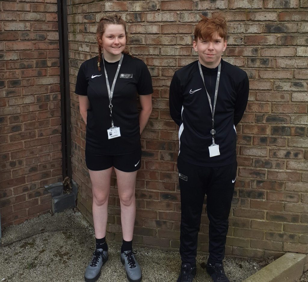Standing side by side, two students dressed in all black sportswear smile looking proudly at us.