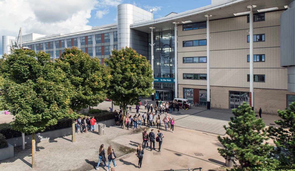 A modern college building named 'The HUB' with a glass entrance, beige brick exterior, and metallic architectural features. The foreground features a landscaped area with green trees and paved walkways. Groups of students are walking and gathering outside the entrance under a blue sky with scattered clouds.