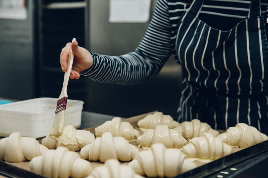 A close up image of a person in chef apron holding a brush, brushing a coating onto pastry croissant shapes