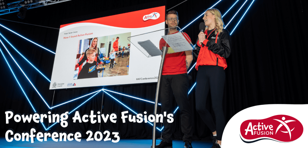 aTwo people in red, black and white sport style uniforms stand on stage in front of a large projection screen. On the screen is a photograph of a young child being coached by an Active Fusion member of staff.