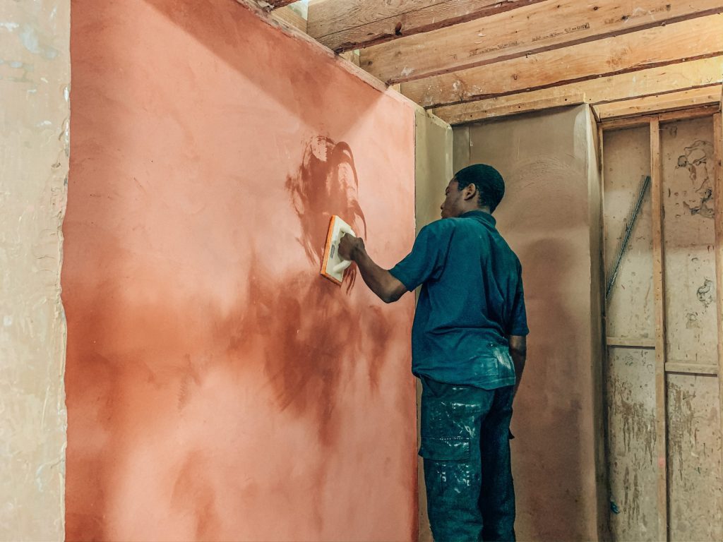 A person stands in front of a wall that they are skimming with plaster. They have a tool in hand as they make the wall smooth