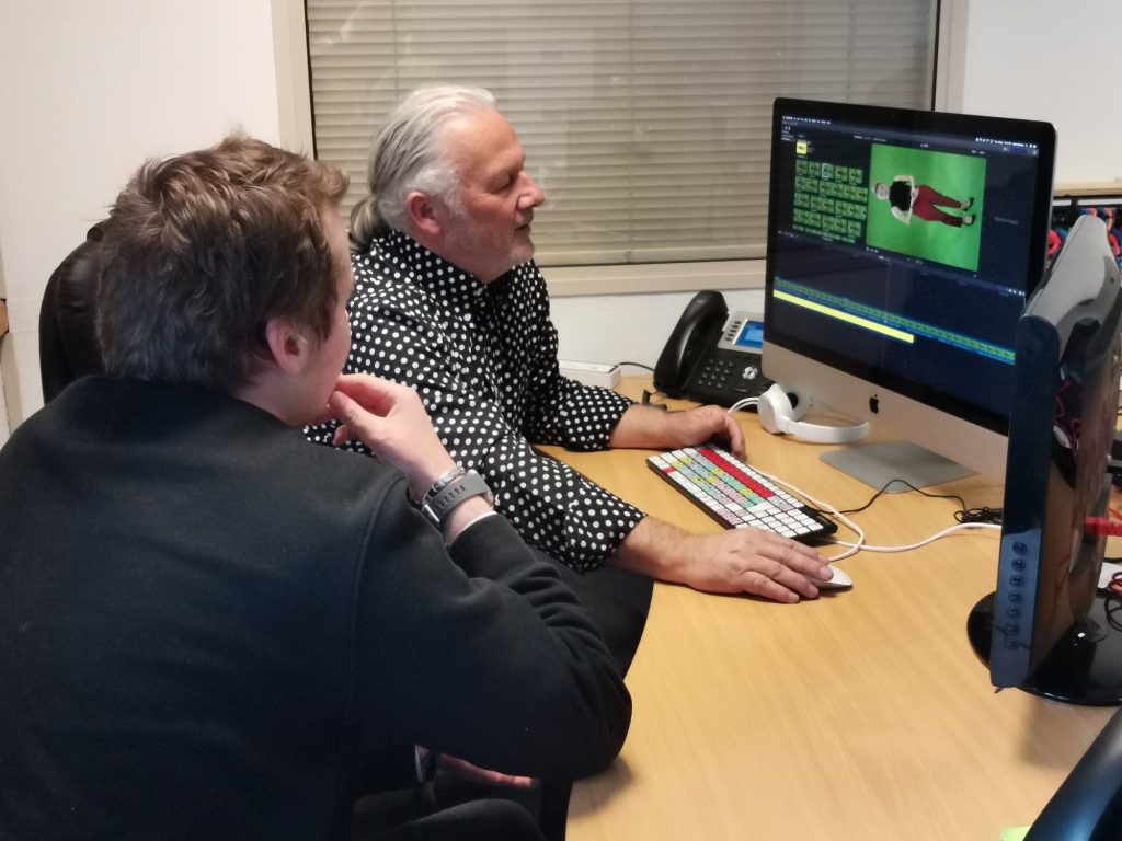 Sat at a desk, two people look at a computer screen. The person furthest, Kevin, is operating the computer which has a photograph of a figure on the screen.