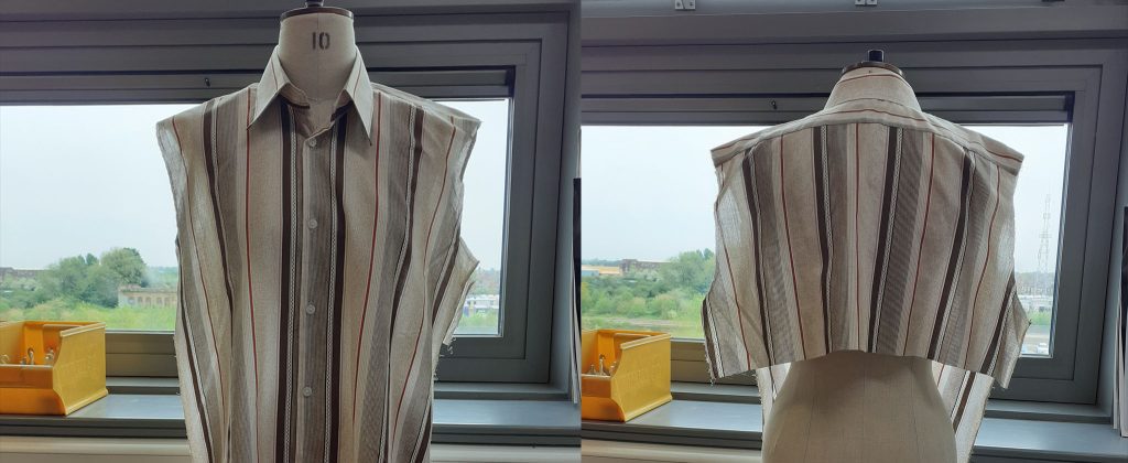 In front of a window, two upper body torso mannequins are dressed in some bibs made from shirts. Each has a vertical striped pattern.