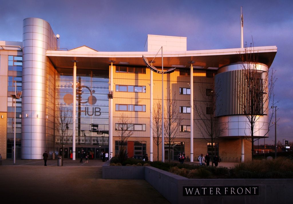 Modern educational building with large glass windows and a metallic curved structure on the right side. The entrance has a glass facade with 'The HUB' logo displayed. The exterior is illuminated by warm sunlight, with people walking near the entrance. A 'WATERFRONT' sign is visible in the foreground, surrounded by landscaped greenery.