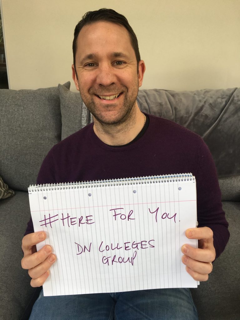 Image featuring Robin Makey sat on a sofa smiling, holding up a sign that says 'Here For You'