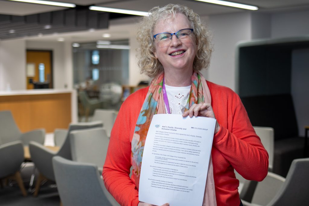 Wearing bright clothing, DN Colleges Group's Karen Dainty smiles proudly at the camera as she holds a document up highlighting the AoC Equity, Divertsity and Inclusion Charter. She stands in a modernly furnished seating area