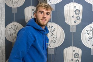 Painting and Decorating student, Henry, stands proudly against a wall he has decorated. Henry wears a blue hoodie flecked with some paint marks, and the wall behind him features an elegant deep blue and white artwork design of many different types of lamps.