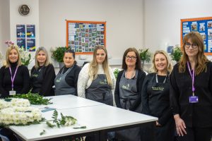 A group of 5 students wearing floristry aprons uniforms stand between two lecturers. In front of the group is a table featuring a floral wreath in the process of being created. Behind the group a variety of floral arrangements in vases can be seen decorating the room