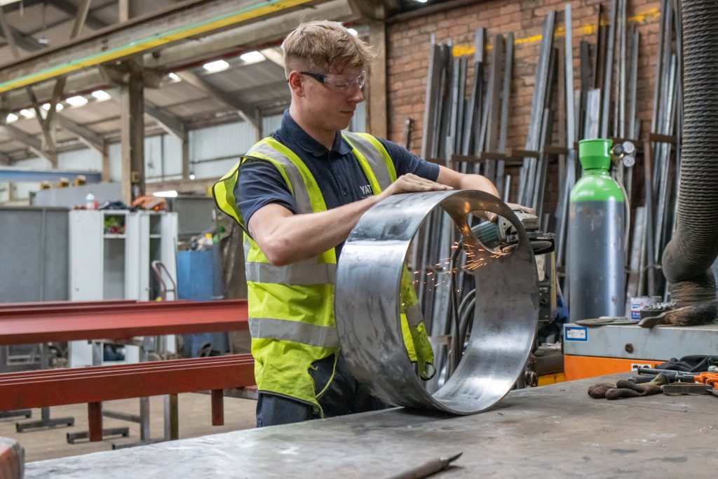 A male apprentice wearing safety goggles and a high-visibility vest works on a metal piece, grinding its edges and producing sparks in an industrial workshop.