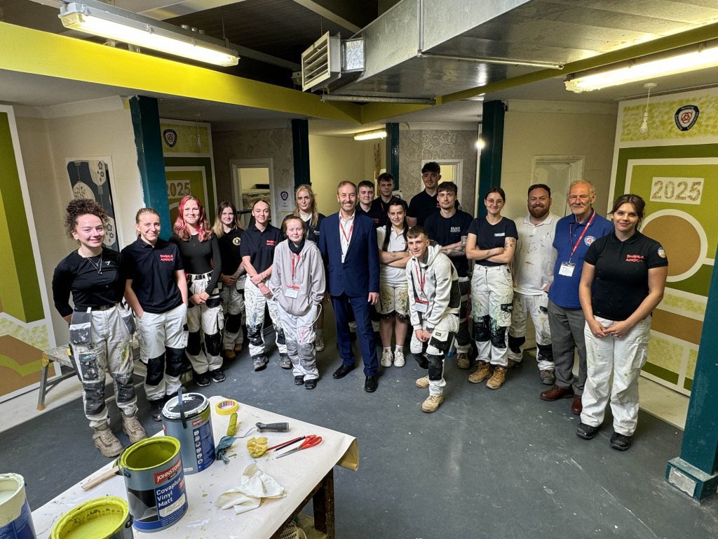 A group of students and instructors stand together in a decorated indoor workspace, posing for a group photo. Most individuals are wearing paint-splattered work trousers and uniforms, suggesting involvement in a painting and decorating project.